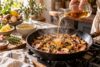 Goldfarbene Flüssigkeit wird aus einer Glaskaraffe in eine heiße Pfanne mit gebratenen Champignons, Schalotten und Thymian gegossen.